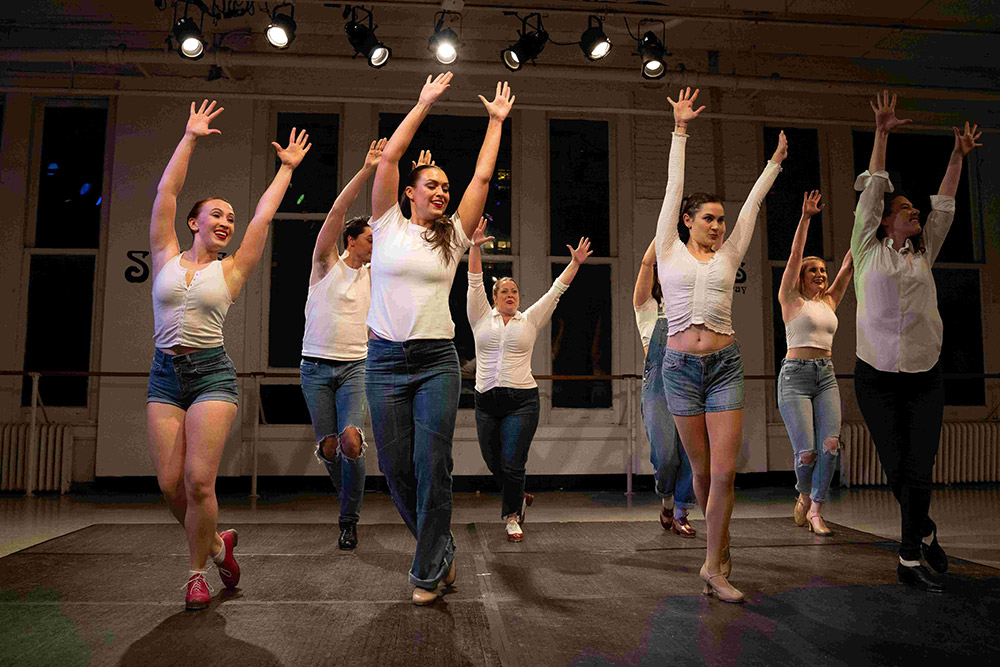 A group of nine people energetically dancing on a stage under warm lighting. They are wearing casual outfits with variations of white tops and denim shorts or jeans. All have their arms raised, creating a joyful and dynamic scene.