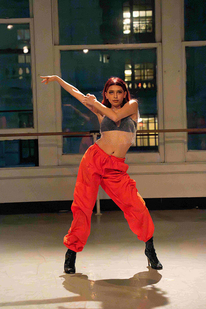 A dancer strikes a dynamic pose in a studio with large windows. They are wearing a mesh top, red pants, and black boots, with one arm extended up and the other bent across their body. The city lights are visible through the windows.