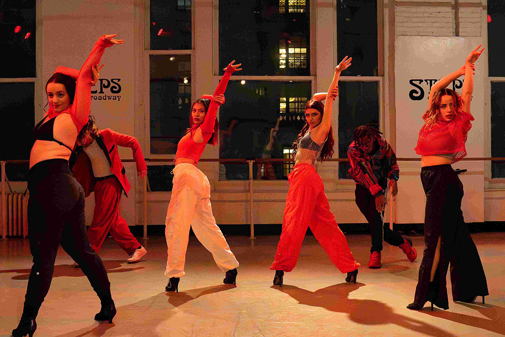 A group of six dancers perform energetically in a studio with large windows. They are dressed in coordinated red and black outfits, striking dynamic poses. The room is softly lit, creating a warm atmosphere.