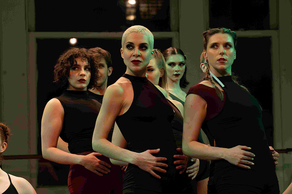 A diverse group of performers in black outfits stand in formation, each with one hand resting on their abdomen. The lighting casts dramatic shadows, and the background shows large windows revealing dim light outside.