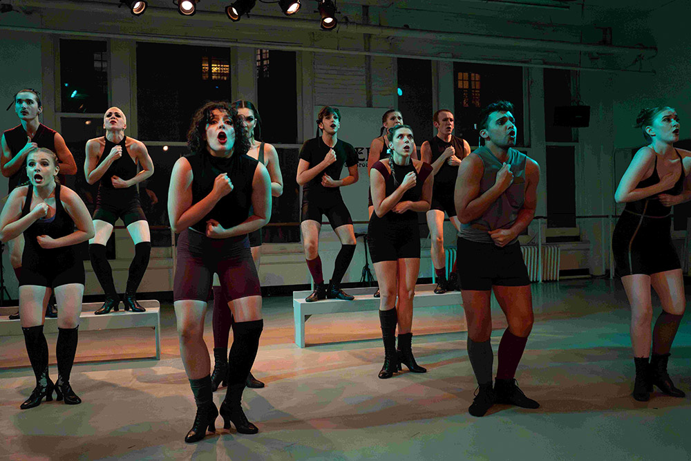 A group of performers in black and dark gray outfits are passionately singing, standing on stage under dramatic lighting. They express intense emotions as they sing with their fists clenched, surrounded by a minimalist set design.