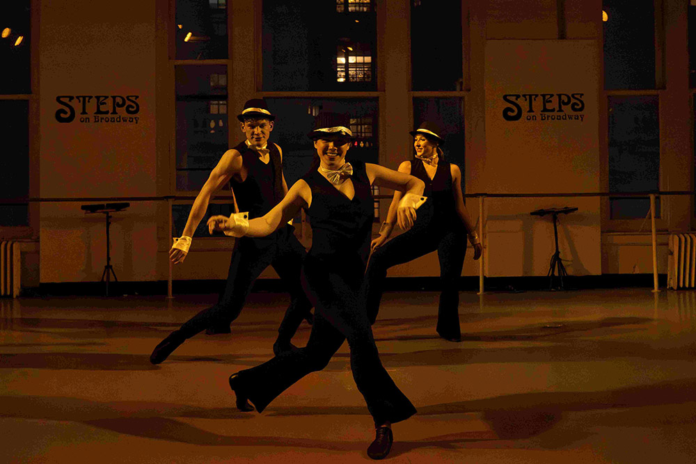 Three dancers in black outfits and hats perform energetically in a dimly lit dance studio with Steps on Broadway signs on the walls. They are mid-movement, showcasing a lively routine.