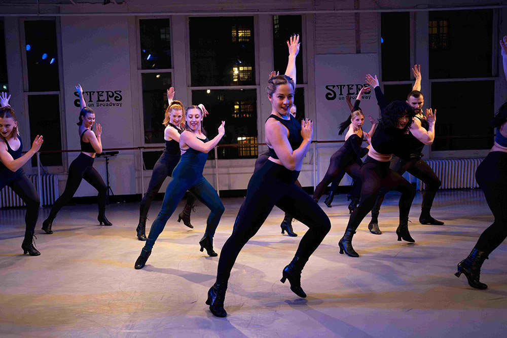 A group of dancers, wearing black outfits and heeled shoes, energetically perform a synchronized dance routine in a studio with Steps logos on the walls. Large windows reveal a nighttime cityscape outside.