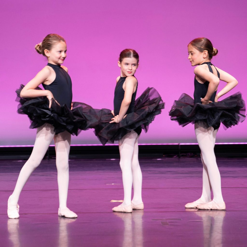 Three Youth Division Ballet dancers in black leotards and black tutus on stage in front of a purple background ready to dance