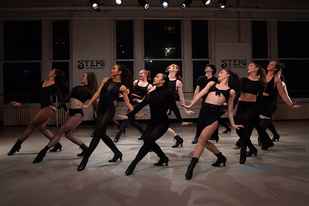 A group of dancers perform in a studio with Steps on Broadway signs in the background. They are wearing black outfits and positioned in a dynamic formation, showcasing expressive poses under bright lighting.
