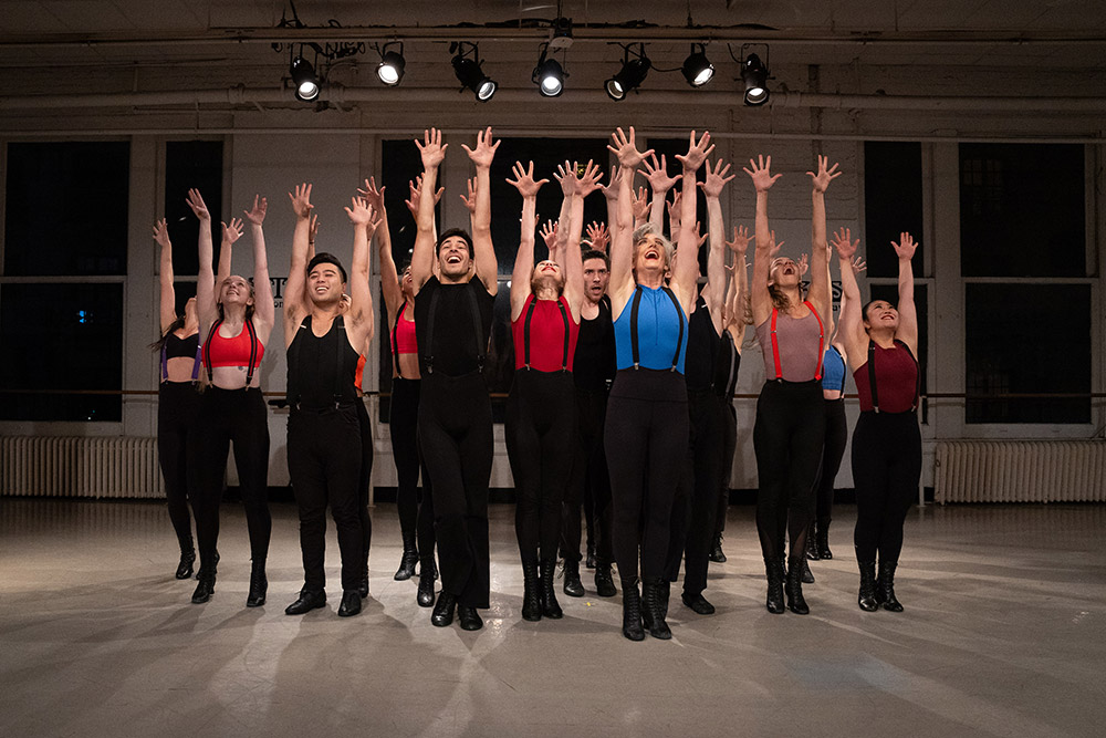 A group of dancers on stage, wearing colorful tops and black pants, perform a routine with arms raised high and legs slightly apart. The background features large windows and overhead lights, creating a dramatic atmosphere.