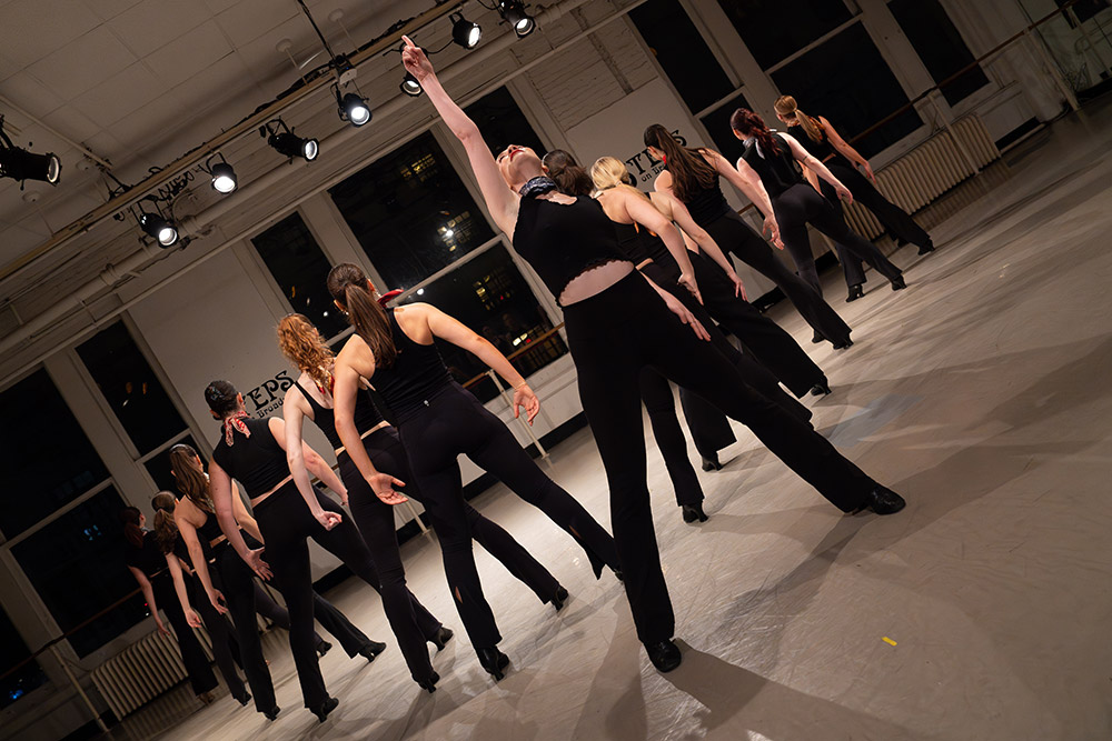 A group of dancers in black outfits performs indoors under stage lights. One dancer in the middle raises an arm triumphantly, while others strike dynamic poses around the room. The scene conveys energy and synchronized movement.