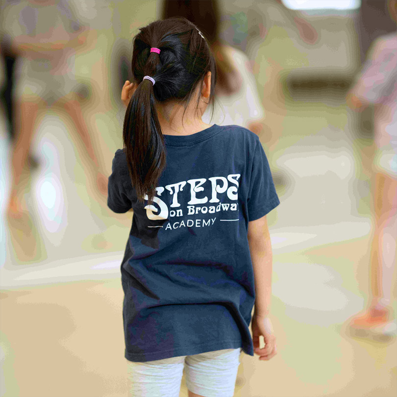 The back of a young steps dancer in a dance studio that is blurred out - her t-shirt has the Steps on Broadway Academy logo on in white on dark blue