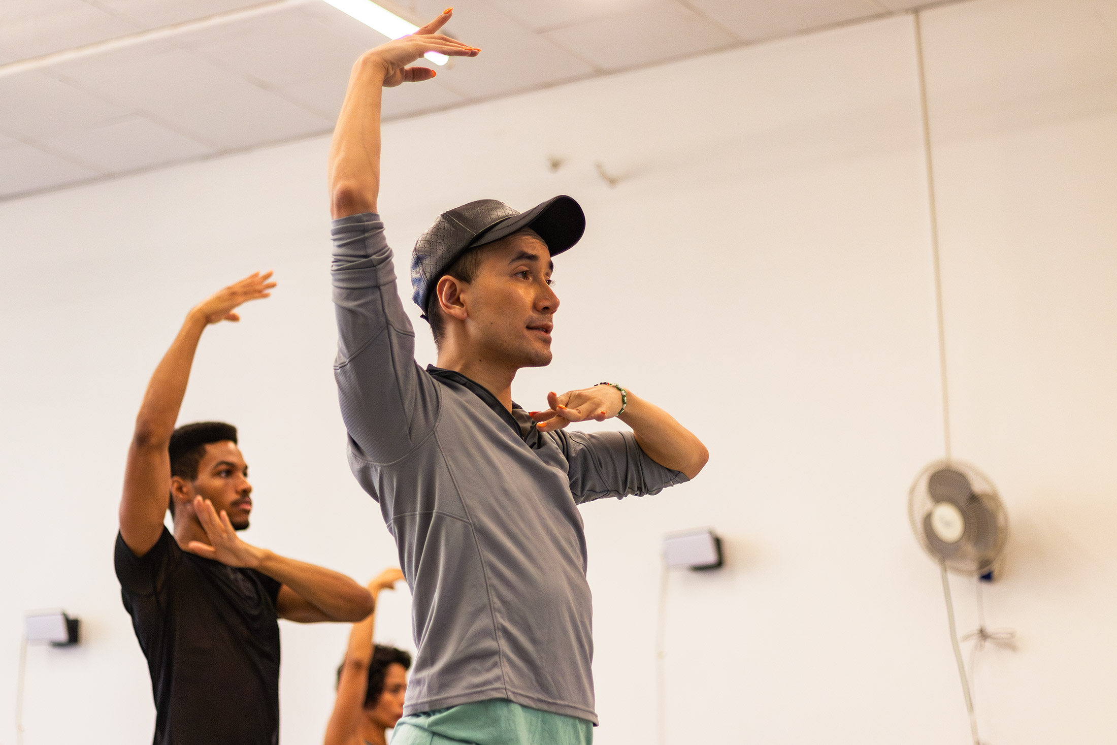 Two people are in a dance studio, practicing with arms raised in graceful poses. One wears a gray shirt and a cap, the other a black shirt. A fan is mounted on the wall in the background.