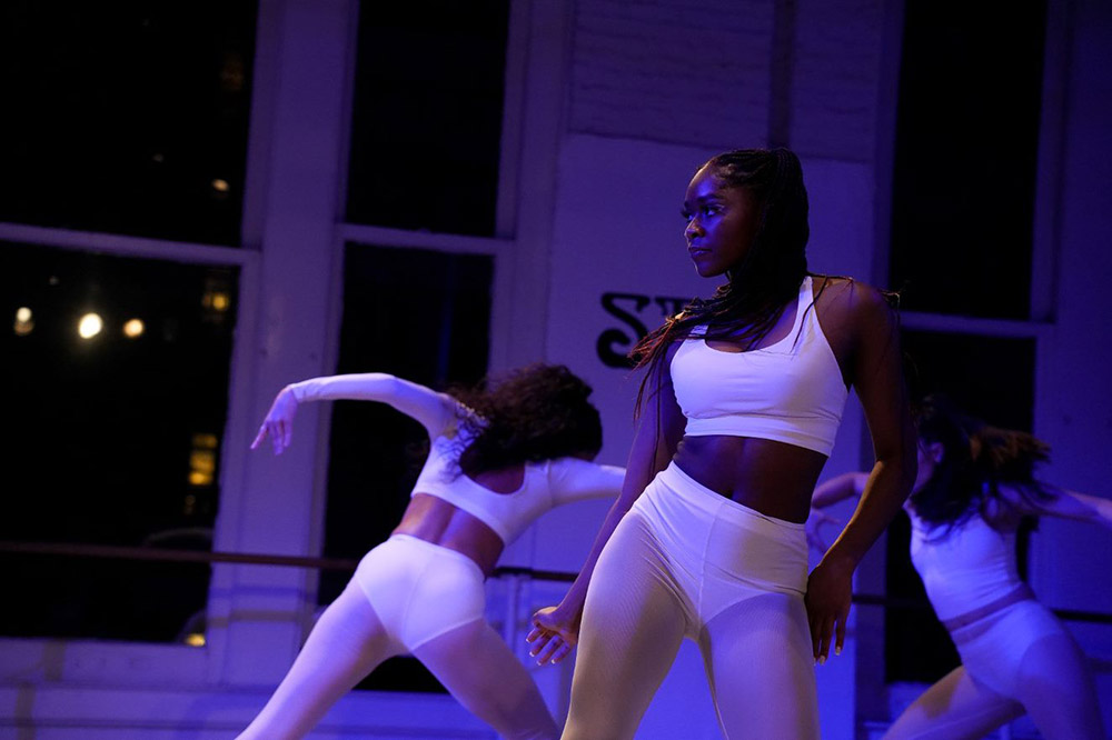 A woman in a white athletic outfit strikes a dynamic pose indoors. Two other women, also in white, dance in the background. The room is dimly lit with blue-purple lighting, and large windows are visible.