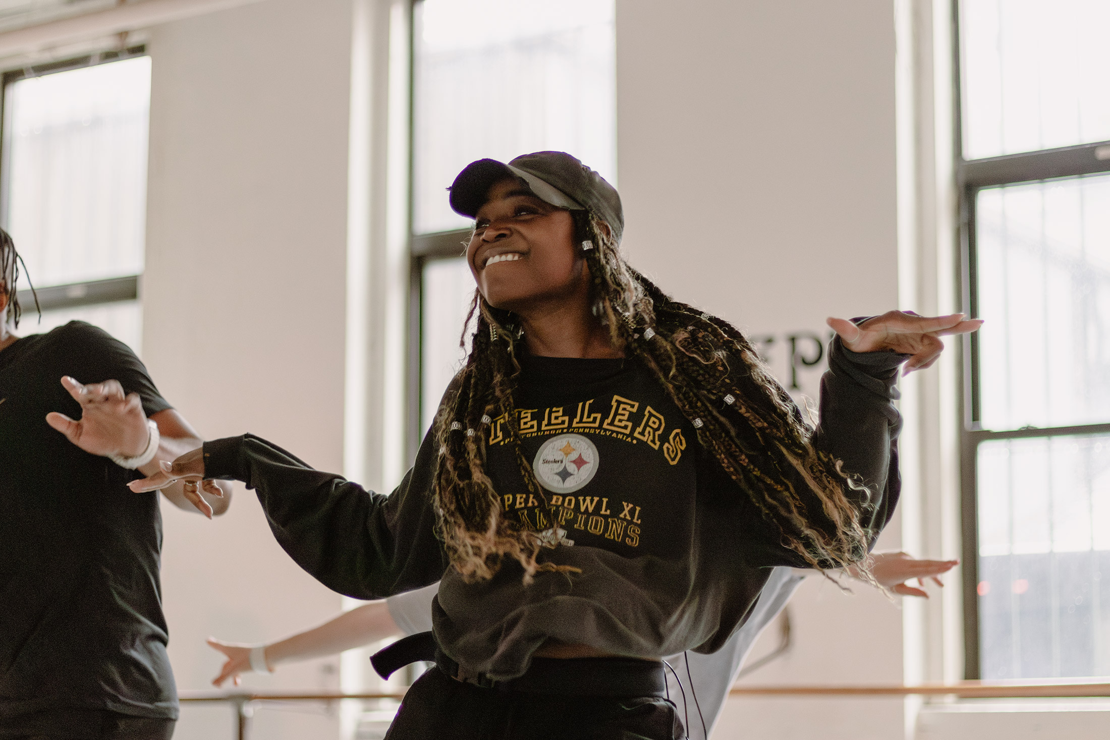 A person dances energetically in a bright room, wearing a cap and a Steelers sweatshirt. Their joyful expression and outstretched arms convey enthusiasm. Large windows in the background allow natural light to fill the space.