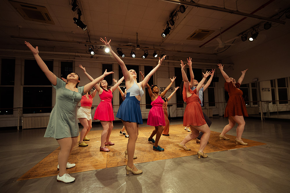 A group of dancers in colorful outfits perform energetically in a brightly lit studio, all with their arms raised and expressions of joy. The background features large windows and stage lighting hanging from the ceiling.