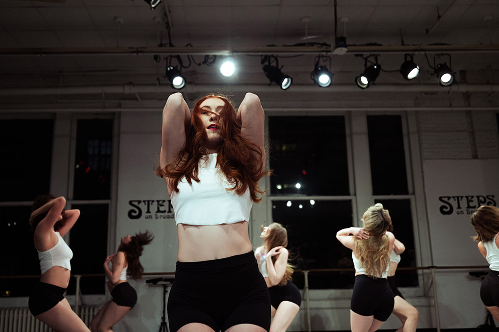 A group of dancers in a studio perform energetically. The focus is on a woman in a white crop top and black shorts, with her hands in her hair. Stage lights illuminate the scene from above. Large windows and a studio mirror are visible in the background.