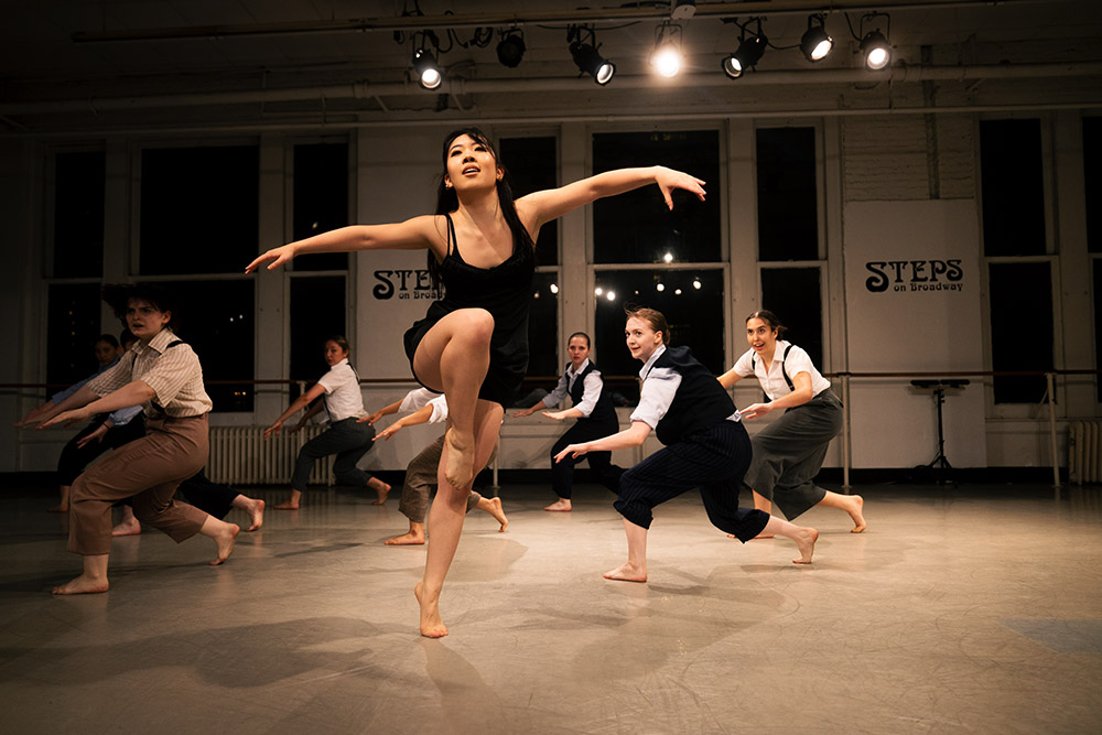 A group of dancers energetically performing in a well-lit studio. The focus is on a dancer in front, leaping gracefully with arms extended. Others follow in dynamic poses. STEPS on Broadway signs are visible on the mirrored walls.