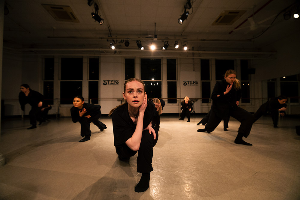 A group of dancers in black outfits perform a synchronized routine in a well-lit studio. The central dancer kneels with one hand on their face, and others are positioned around them in dynamic poses. Looming spotlights create dramatic shadows on the floor.