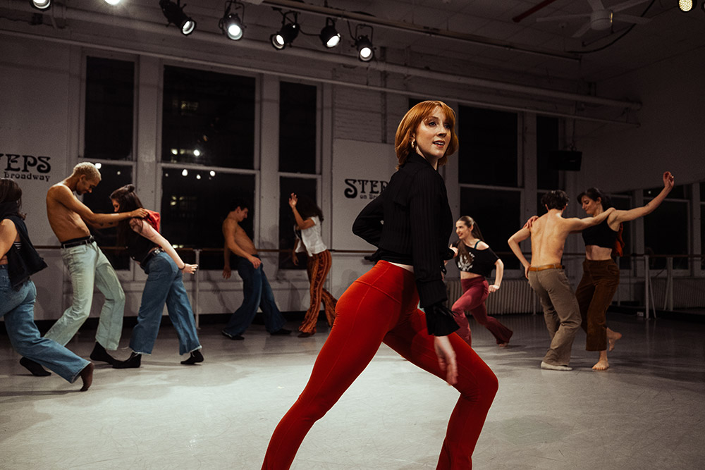 A group of dancers is energetically rehearsing in a dance studio with large windows, under bright overhead lights. They are wearing casual dance attire, and one dancer in red pants is prominently featured in a dynamic pose in the foreground.