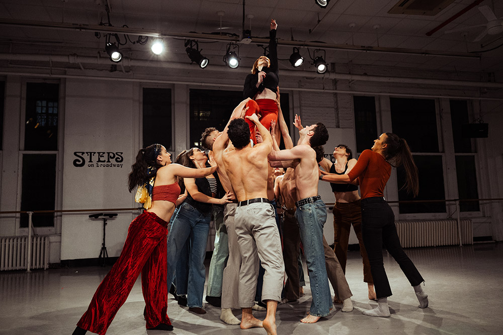 A group of dancers in a studio, with one dancer elevated in the center. They reach upwards, while others surround and support her. The room is well-lit with a large mirror and the words STEPS on Broadway on the wall.