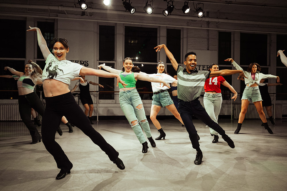 A lively group of dancers perform energetically in a studio, extending their arms and legs in unison. They are dressed in casual attire, and the room is brightly lit with spotlights.