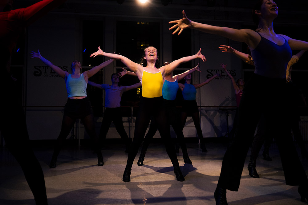 A group of dancers in a studio joyfully stretching their arms wide, wearing colorful tops under dim lighting. The central dancer in a yellow top stands out.