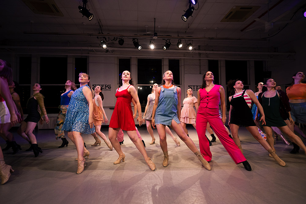 A diverse group of dancers perform energetically on stage, each striking a pose. They wear vibrant outfits, with the central dancer in red. Bright stage lights illuminate the scene, creating a lively and dynamic atmosphere.