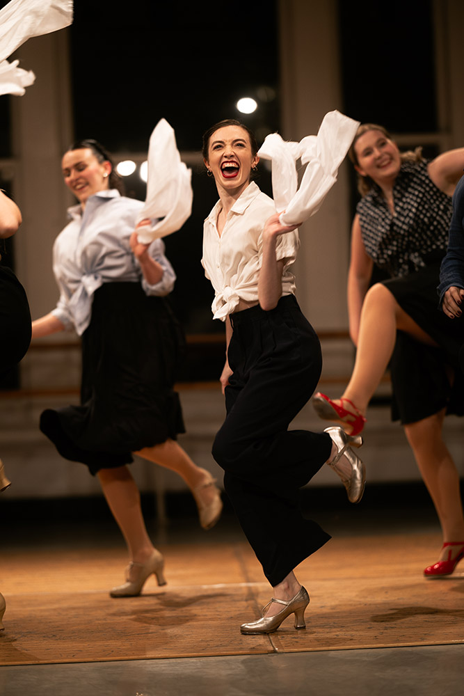 A group of women energetically dancing on stage, wearing vintage-style clothing with black skirts, white blouses, and heels. The central dancer is smiling broadly, swinging a white scarf, and captured mid-step.
