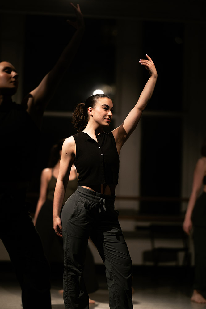 A dancer poses gracefully with one arm extended upwards in a dimly lit studio. She wears a sleeveless black top and dark pants, surrounded by other dancers in the background. The atmosphere is focused and calm.