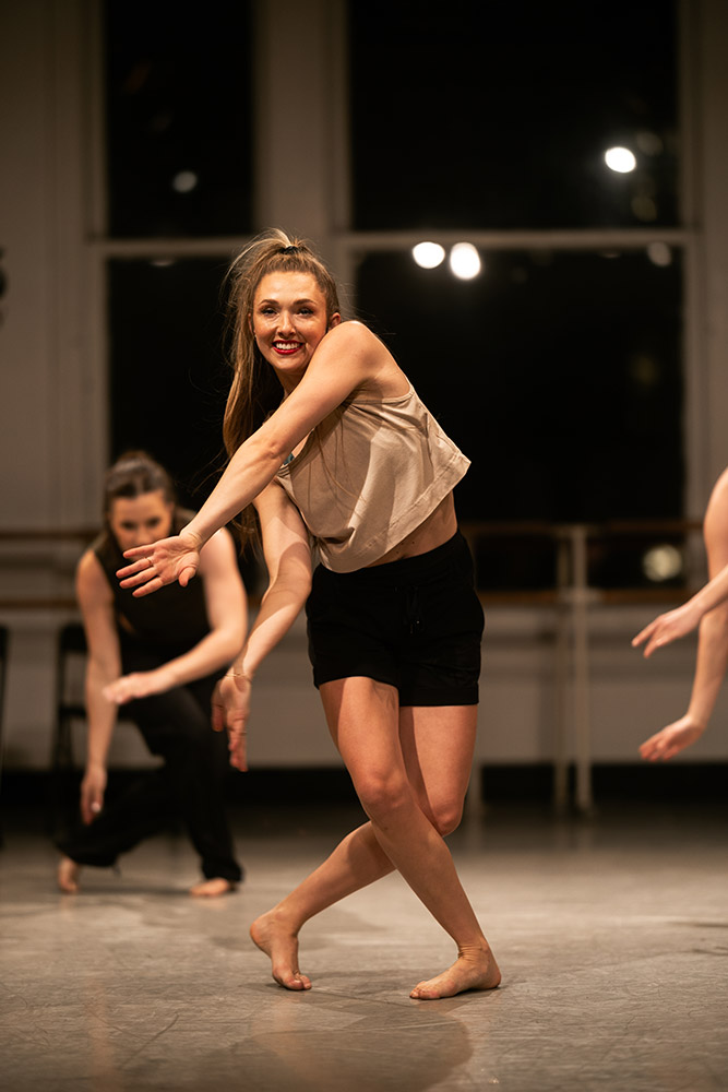 A woman in a beige top and black shorts joyfully dances in a studio with large windows. She is surrounded by other dancers who are partially visible, all immersed in the choreography and expressing themselves through movement.