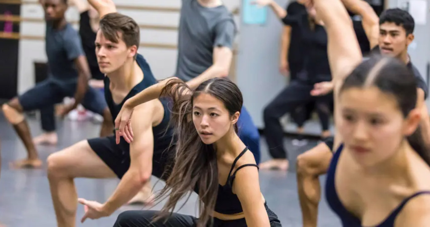 A group of dancers rehearses in a studio, focusing on a contemporary routine. Theyre dressed in athletic wear, striking dynamic poses. The room has mirrors and barre equipment, creating a vibrant and intense atmosphere.