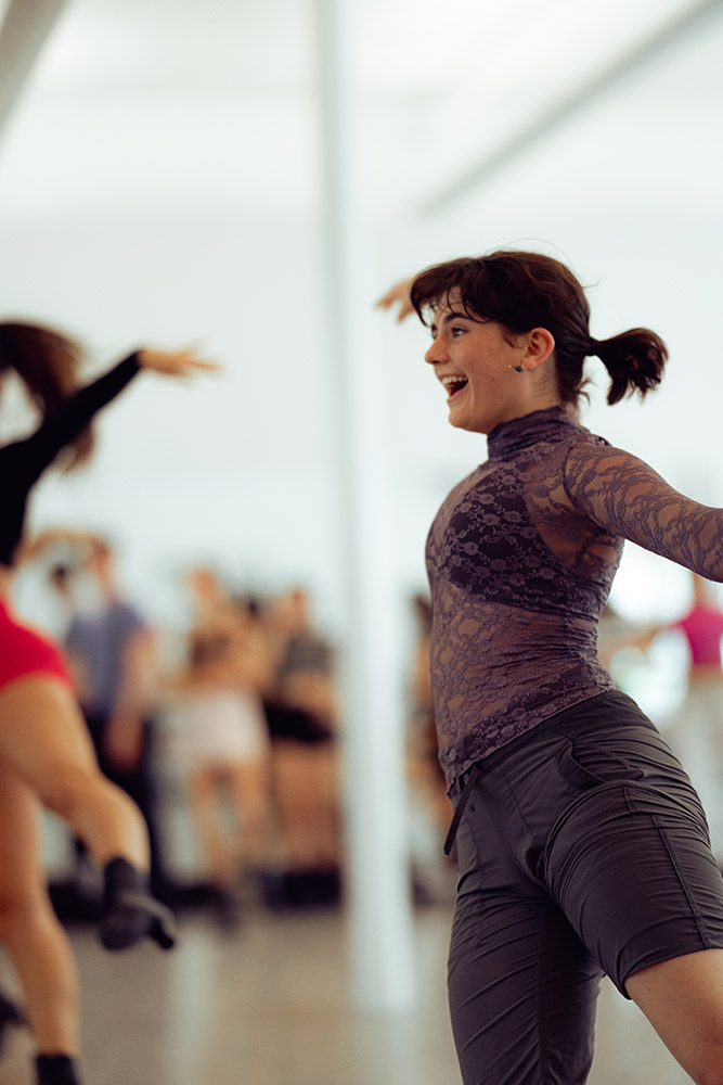 A person with brown hair is joyfully dancing in a studio, wearing a purple lace top and gray pants. Others in colorful outfits are dancing in the background. The atmosphere is lively and energetic.