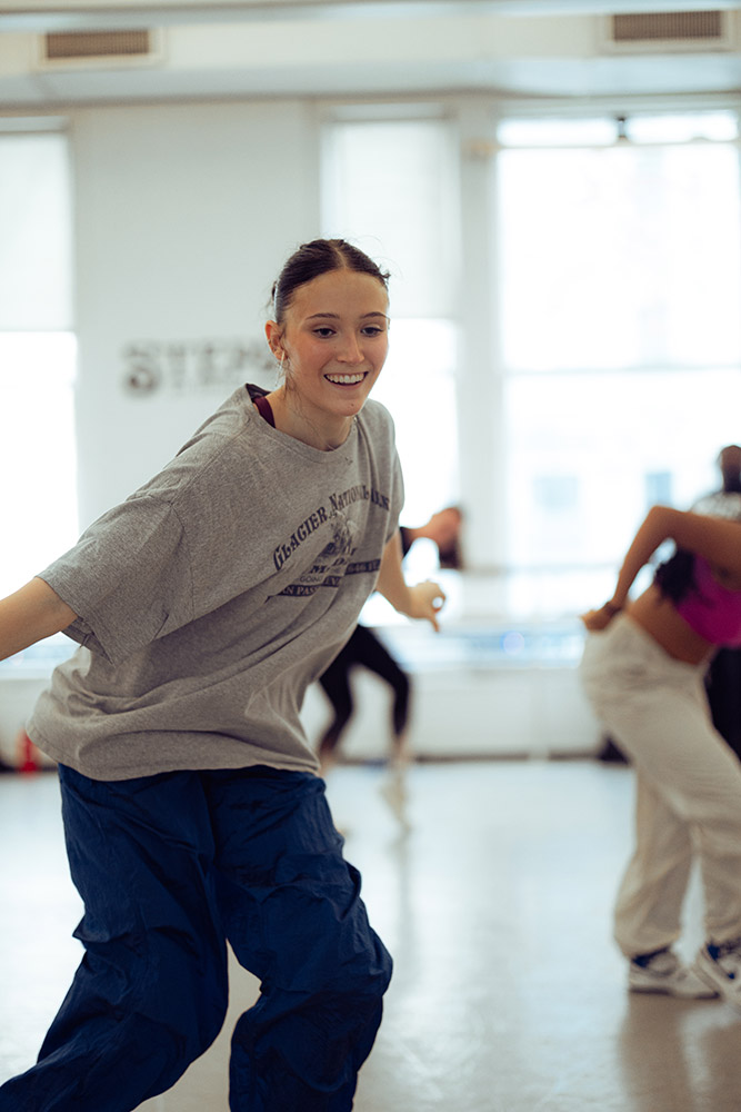 A person in a grey t-shirt and blue pants smiles while dancing in a studio with sunlit windows. Other dancers are blurred in the background.
