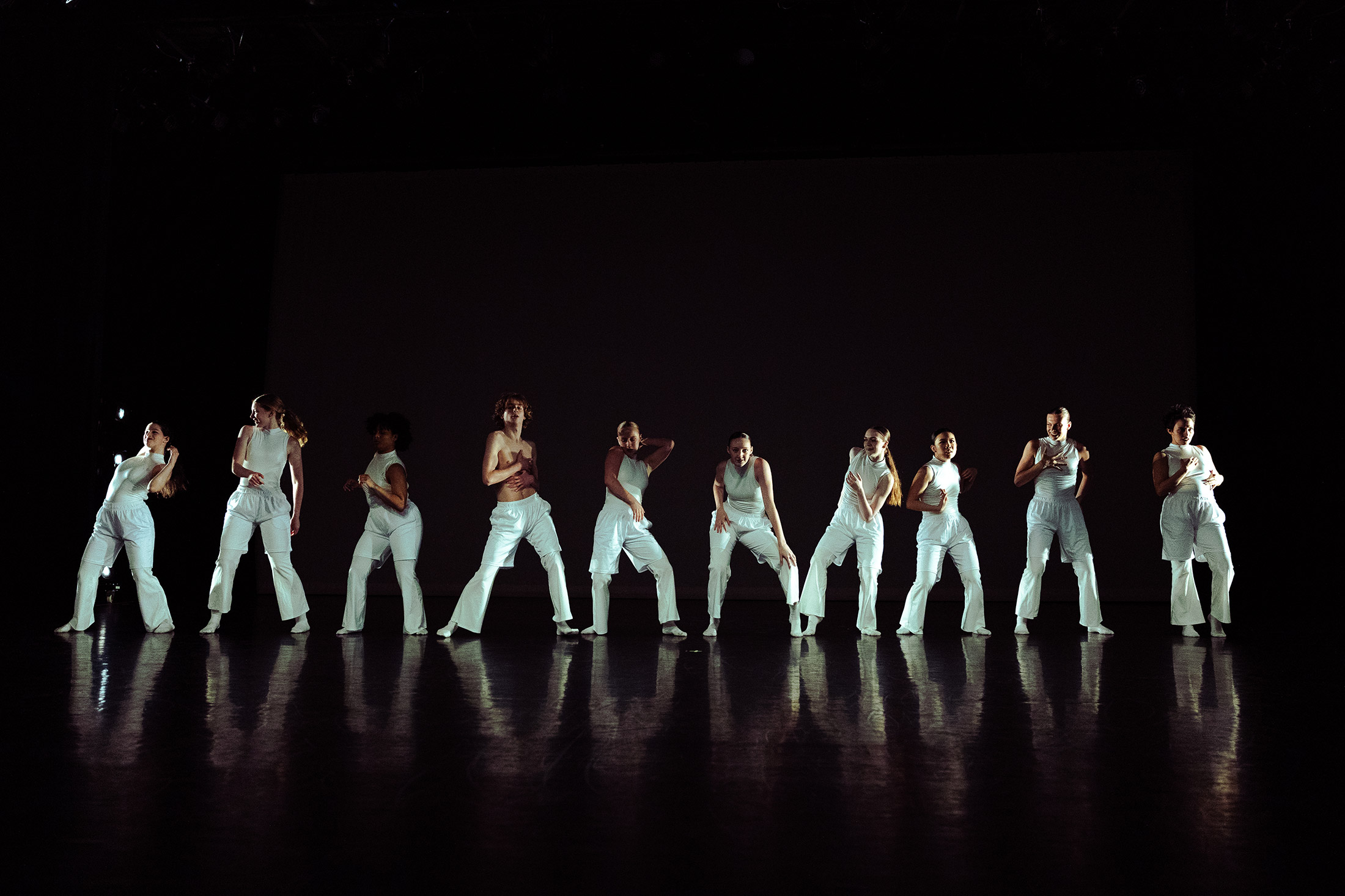 A group of dancers in white outfits perform on a dimly lit stage, striking a pose in a row. The background is dark, and the floor reflects their silhouettes, creating a dramatic scene.