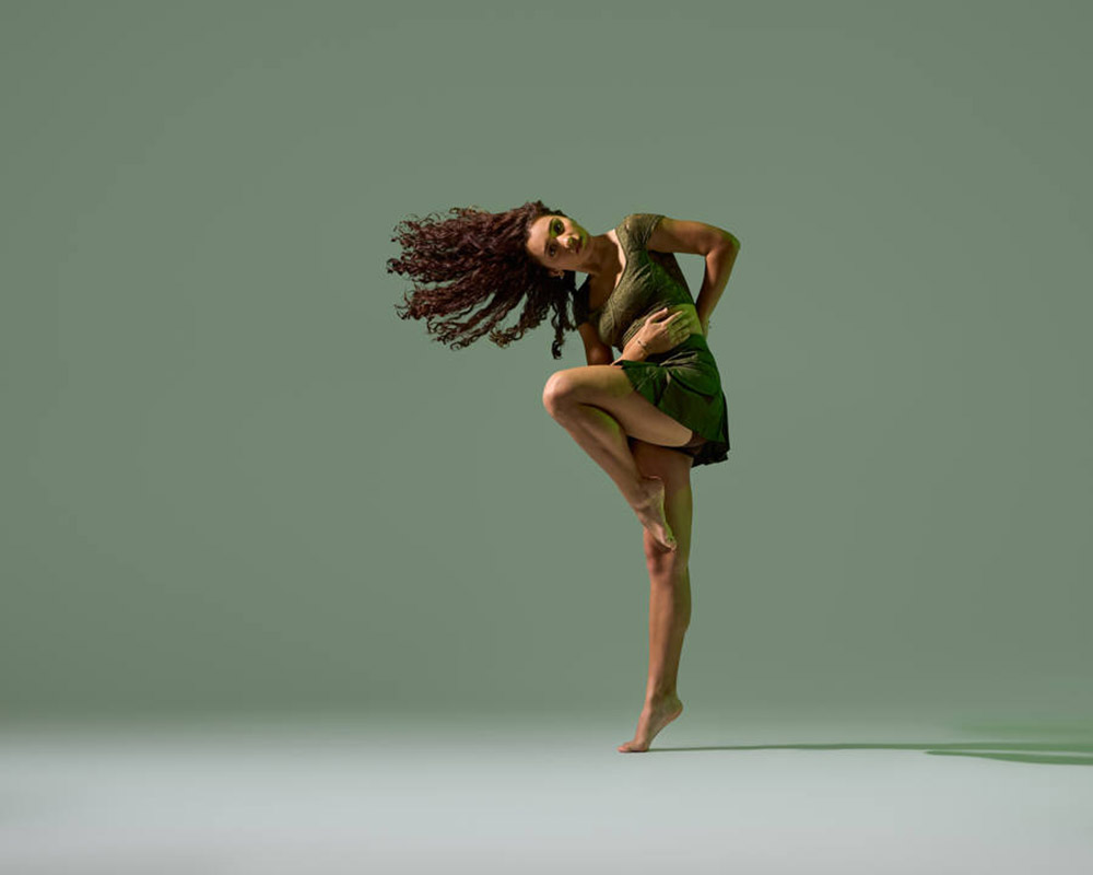 A dancer with curly hair performs an expressive pose on one leg, wearing a green dress against a matching green background. Her arms are wrapped around her lifted knee, and her other leg is extended gracefully.