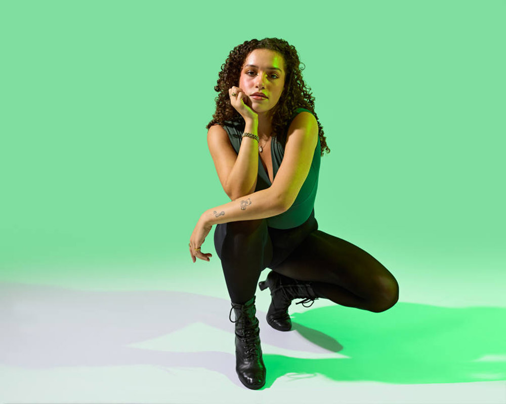 A person with curly hair kneels on one knee against a green background. They are wearing a dark sleeveless top, black leggings, and black lace-up boots, resting their chin on their hand.