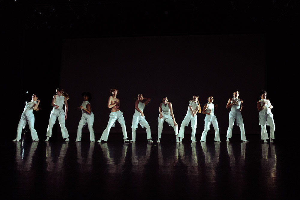 A group of dancers in white outfits perform on a dark stage. They are arranged in a line, each striking a pose with one leg bent and arms in various positions. The lighting casts a dramatic shadow behind them.