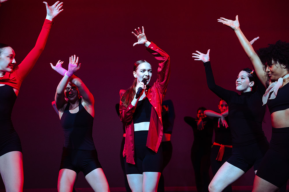 A woman in a red jacket sings into a microphone on stage, surrounded by dancers in black outfits striking dynamic poses with arms raised, under a red spotlight.