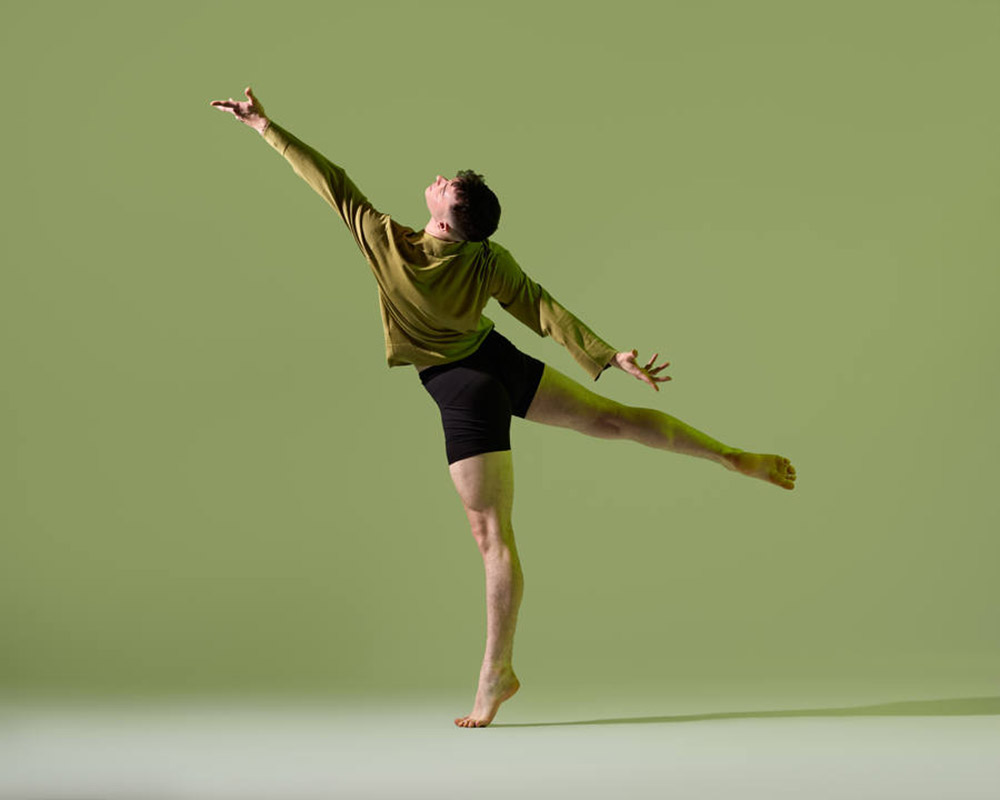 A dancer performs a graceful pose on one foot, with one arm raised and the other extended to the side. They are wearing a green long-sleeve shirt and black shorts, against a light green background.