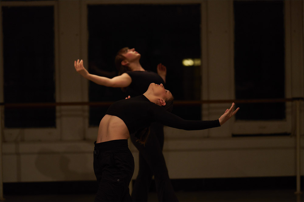 Two dancers in black outfits are gracefully arching backwards with arms extended, in a dimly lit studio. The focus is on their expressive movement, set against large windows in the background.