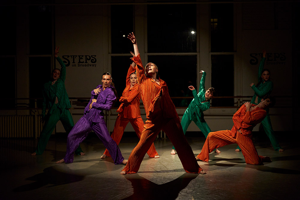 A group of six dancers wearing vibrant costumes in orange, purple, and green perform energetically in a studio. Their expressive poses are captured under dramatic lighting, with a STEPS on Broadway sign visible in the background.