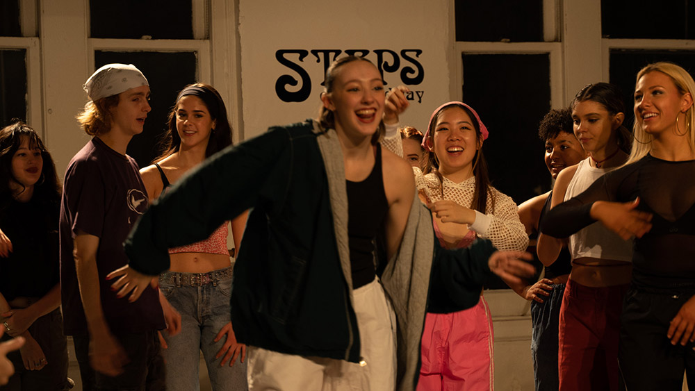 A group of smiling young adults, casually dressed, gather in a dance studio with Steps on Broadway on the wall. One person in a green jacket is in the foreground, animated and cheerful, as others look on encouragingly.
