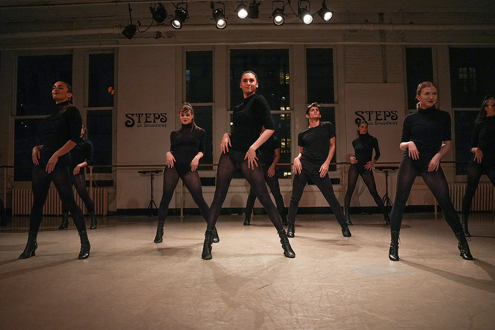 A group of dancers in black outfits and heels perform a synchronized routine in a studio with large windows. The room is dimly lit with spotlights above, and STEPS signs are visible on the walls.