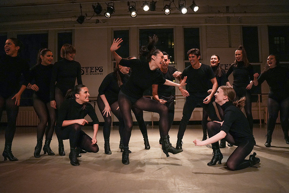 A group of dancers wearing black outfits and high-heeled boots perform energetically in a studio with STEPS written on the wall. They appear joyful and engaged, with some crouching and others leaping mid-dance.