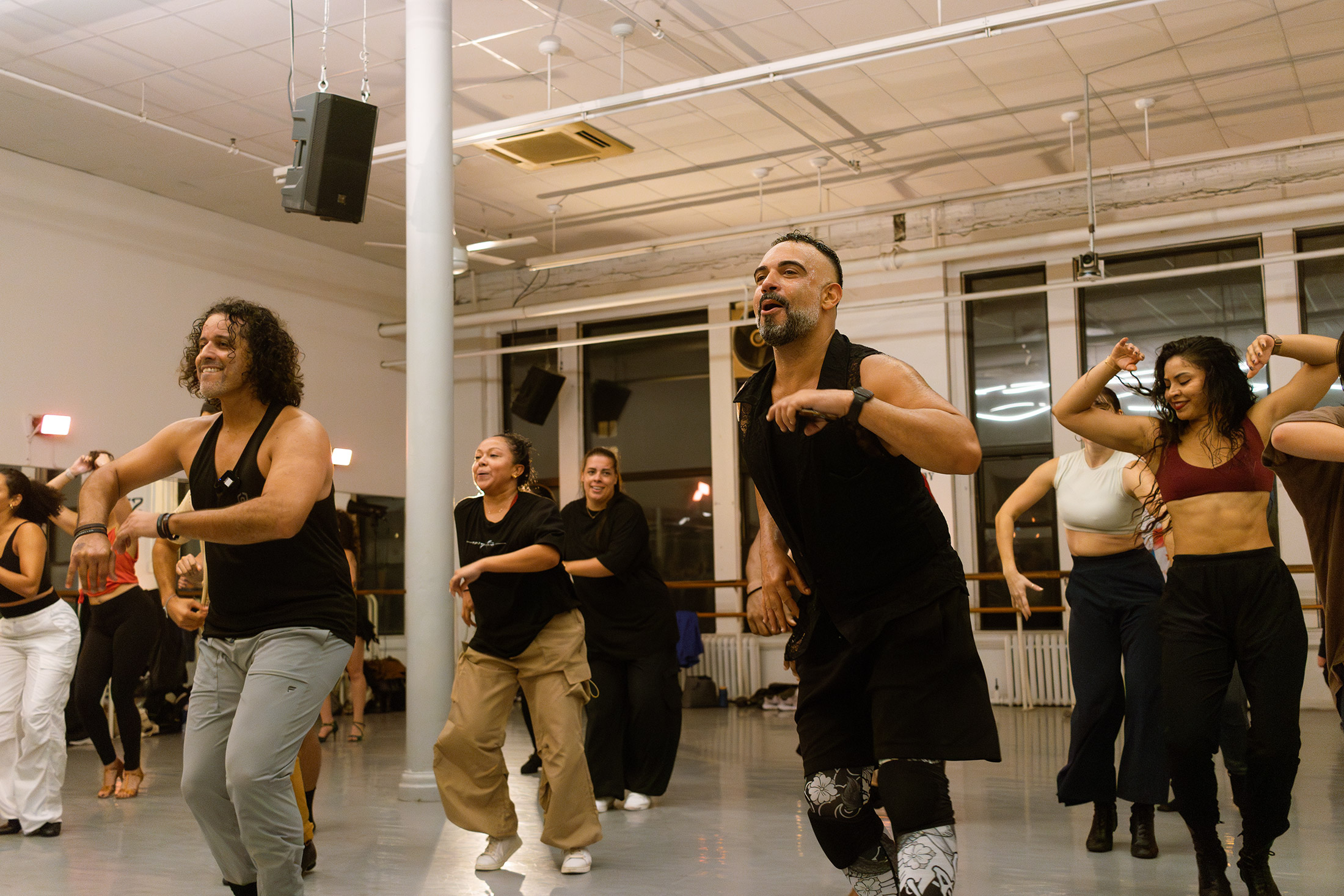 A diverse group of people are dancing energetically in a studio with large windows. The room has a bright, lively atmosphere, and the dancers appear joyful and engaged in their movements.