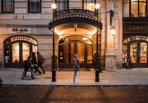 A street scene showing the entrance of Hotel Belleclaire at dusk, with warm lights illuminating the facade. People walk by on the sidewalk, and the shops White + Warren and LUSH are visible alongside the hotel entrance. Autumn leaves are on the ground.