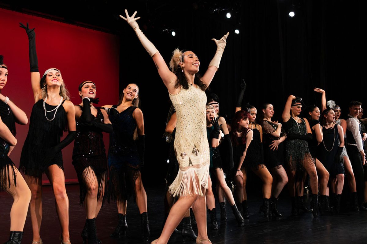Steps IAP dance students on stage- the from woman in a white flapper style dress facing stage left, arms raised in joy