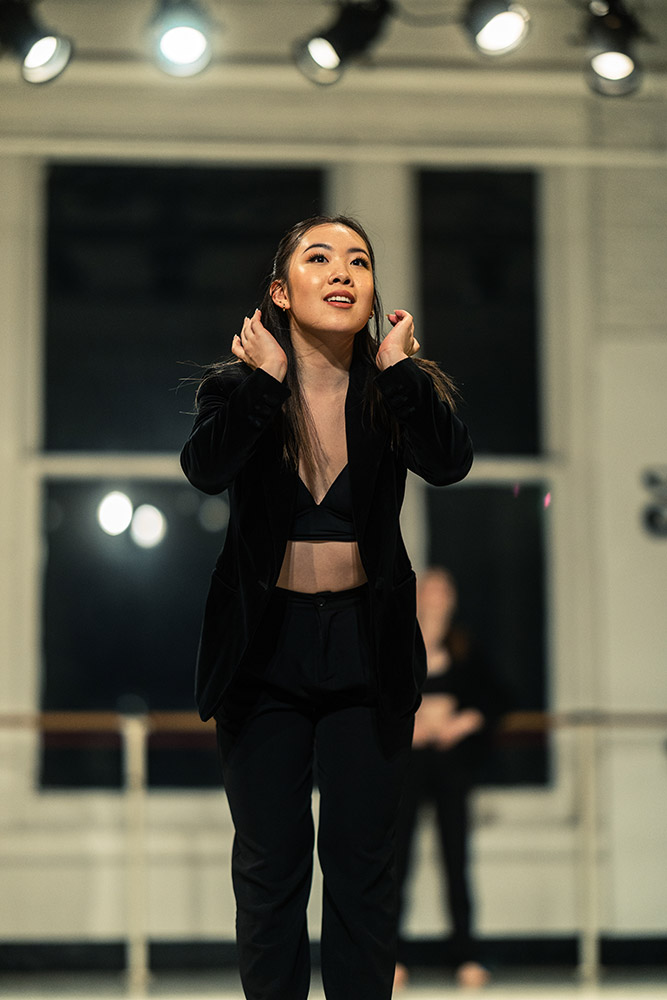 A woman in a black outfit performs a dance routine on stage, with soft lighting illuminating her. She is holding her hair back, and large windows are visible in the background.