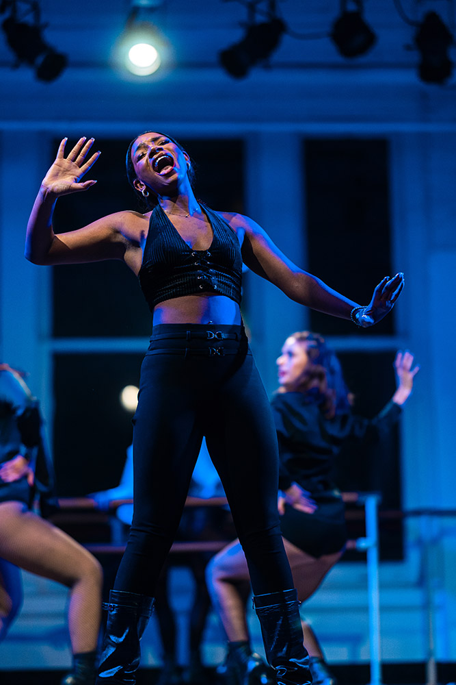 A performer in a black outfit energetically sings on stage, with their mouth open and hand raised. They stand under blue stage lighting, with a blurred background of other performers.