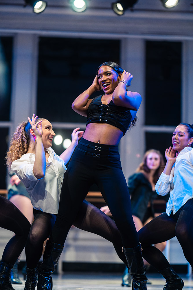 A group of dancers performing energetically on stage. The central dancer, wearing a black outfit, is smiling and leading the routine. Other dancers, dressed in white tops and black pants, are enthusiastically following the choreography. Stage lights illuminate the scene.