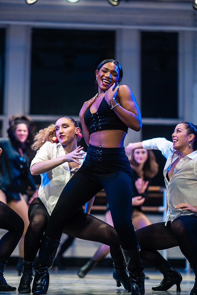 A group of dancers performs energetically on stage. The lead dancer wears a black crop top and pants, with others in the background dressed in coordinating outfits. The scene is dynamic and joyful, under bright stage lights.