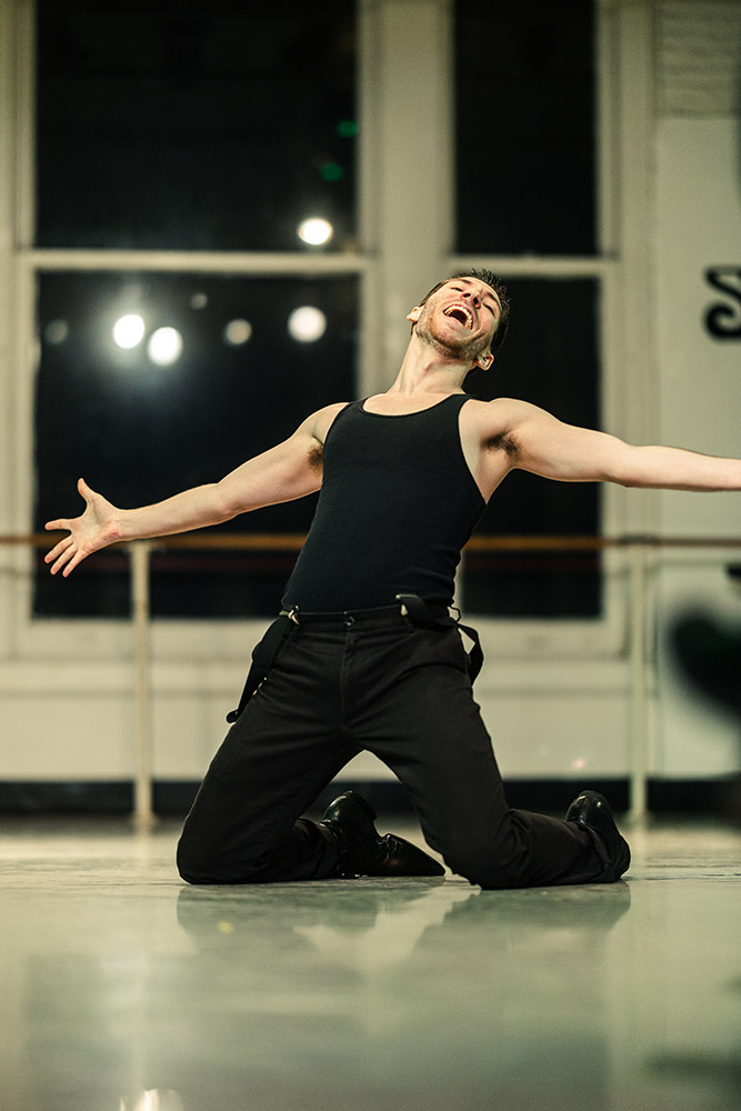 A person in a black tank top and pants energetically kneels on a stage with arms wide open, appearing to sing or shout, in a brightly lit dance studio.