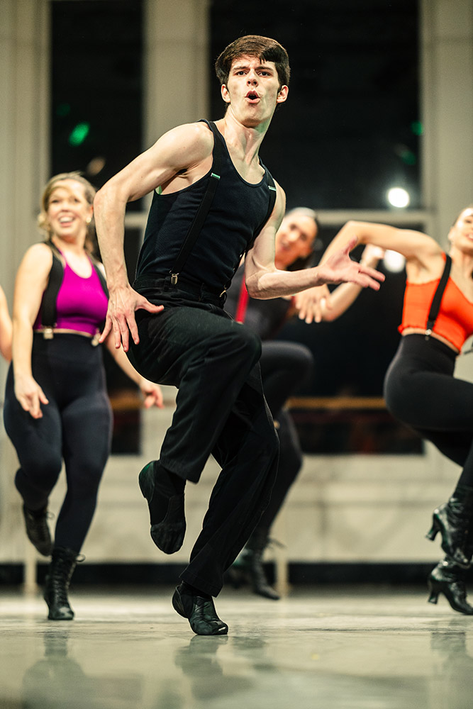 Dancers energetically perform in a studio, led by a male dancer in a black tank top and pants. Other dancers, wearing brightly colored tops, move in the background. The room has large windows, and the atmosphere is dynamic and lively.